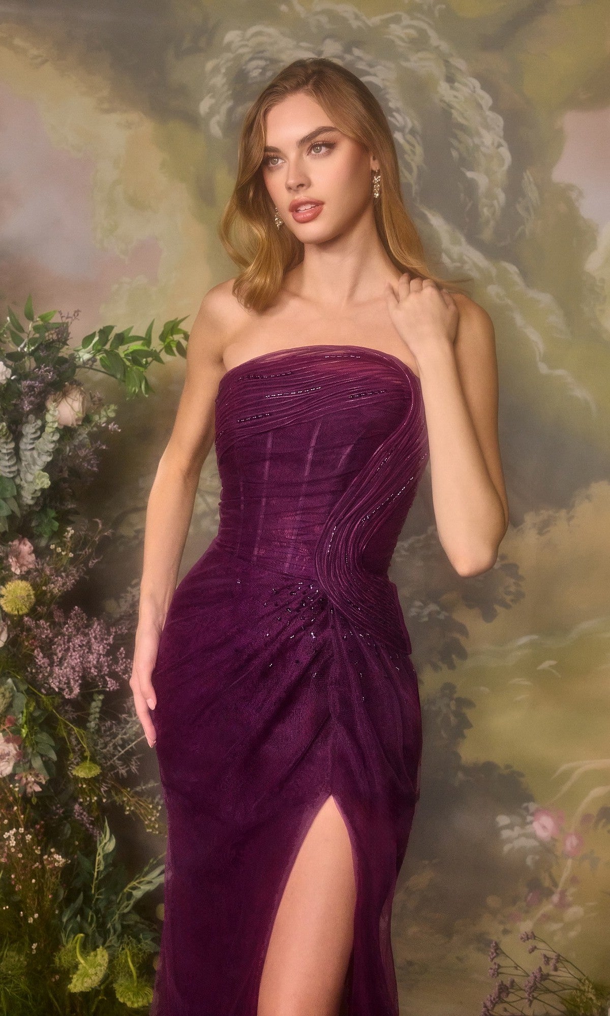 A woman stands before a floral and painted backdrop wearing the Andrea and Leo A1497 long prom dress, a strapless deep purple gown featuring a thigh-high slit.